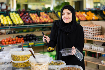 Pleased woman in sheila puts pickled olives into a container in the grocery section of a supermarket