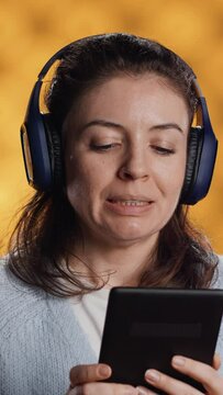 Vertical video Woman wearing headphones, practicing new language vocabulary using online app on tablet, isolated over studio background. Lady speaking foreign phrases, reading them on device screen