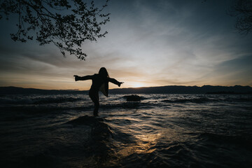 A silhouette of a young girl with arms outstretched, standing in a lake against a vivid sunset,...