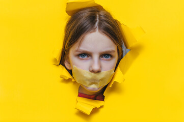 A child, his mouth covered with masking tape, peers through a torn hole in yellow paper. The...
