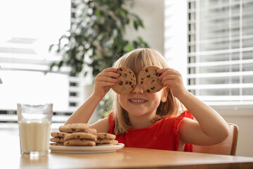 Joyful girl with cookies over eyes, having a delightful snack time with milk and cookies. 