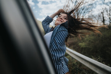 A joyful young woman with her hair blowing wildly in the wind sitting on car window, embodying a...