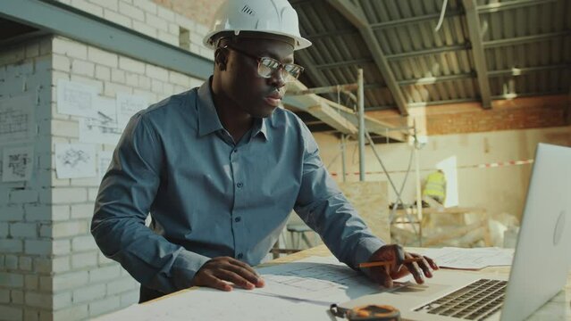 African American male architect wearing hardhat sketching floor plan on paper and working on laptop during the day at construction site