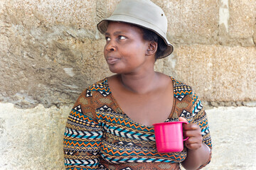 young african chubby woman with a hat holding a mug in the poor township, informal settlement