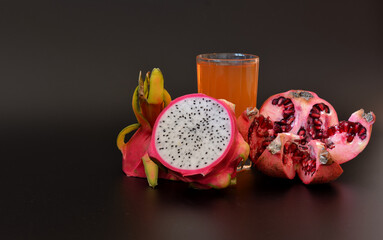 A glass of freshly squeezed fruit juice on a black background, next to pieces of ripe pitaya and a pomegranate fruit with seeds.
