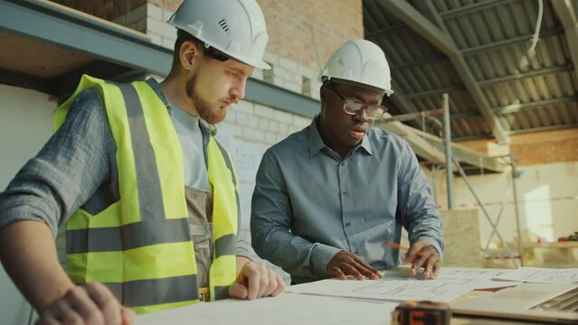 African American foreman describing floor plans on paper to male Caucasian building contractor in safety vest and hardhat when working together at construction site