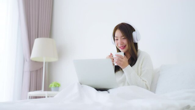Morning Time in Bedroom Young Asian Woman Happy Drinking Milk and Working on Computer Laptop, Wearing headset, Sitting on Bed at House