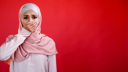 Omg embarrassment. Shame fear. Keep quiet. Overwhelmed frightened sad woman in hijab covering mouth with hand isolated on red empty space background.
