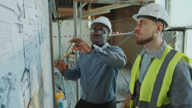 African American male architect in hardhat and formal shirt pointing at floor plans on wall and giving instructions to Caucasian building contractor at construction site