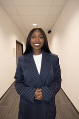 Captured in a corporate hallway, this image features a Black woman wearing a sharp pinstripe suit with a radiant smile. Her professional attire and poised demeanor convey confidence and success within