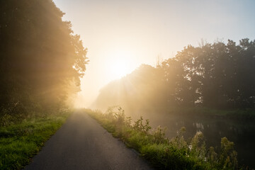 The image portrays a secluded pathway alongside a river, with a radiant sunburst piercing through...