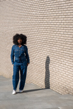 A woman stands casually against a pale brick wall, her face accidentally obscured by a watermark in the image. She's dressed in a stylish denim jumpsuit paired with white sneakers, embodying a relaxed
