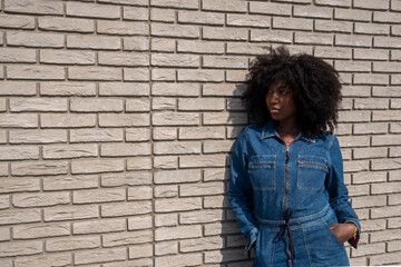 This candid photo showcases a young Black woman with a striking afro hairstyle, dressed in a trendy denim jumpsuit. She leans against a white brick wall, her stance exuding a relaxed yet confident