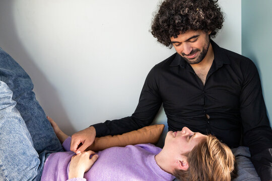 This warm photograph captures an intimate moment between a couple in the comfort of their home. A man with curly hair and a black shirt smiles fondly as he looks down at his partner, who lies relaxed