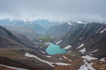 Most beautiful turquoise alpine lake in stony rocky green valley against mountain range silhouette in rainy low clouds in gray sky. Large snowy mountains in light transparent mist in rain bad weather. - 792333456