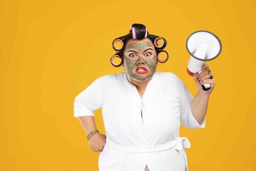 Portrait of a fat housewife wearing a natural mask and holding a megaphone isolated on a yellow...
