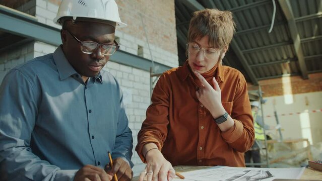 Young Caucasian woman discussing floor plan on paper with African American building contractor while visiting construction site
