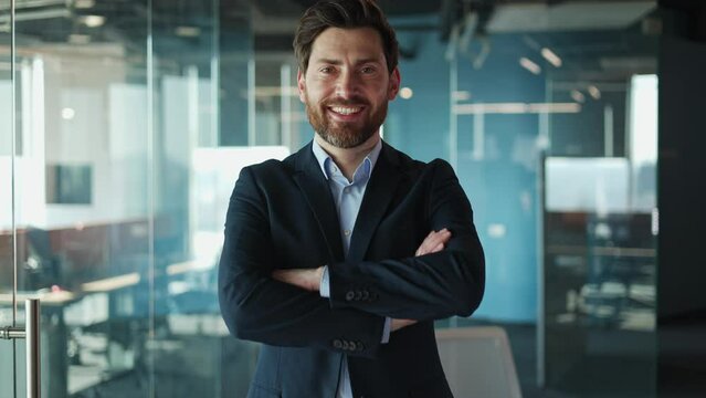 Bearded caucasian man standing confidently with arms crossed against glassy backdrop of corporate office wall. Male cheerfully gazing in camera and portraying determination and professionalism.