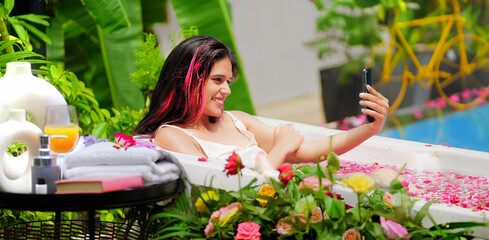 Indian Asian smiling gen z young adult girl sitting rose petal tub take selfie use mobile cam hold...