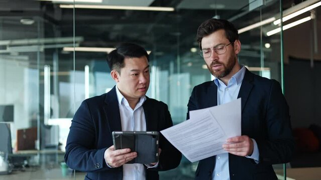 Confident males tapping on digital tablet while concentrated partner reading important documents in modern office interior. Busy diverse men checking sameness of online and printed form indoors.