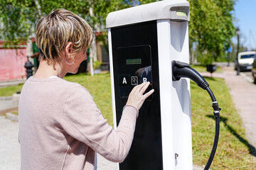 Woman uses touchscreen on EV charging station, selecting options for her car.