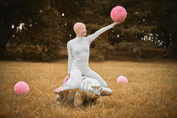 Young hairless girl with alopecia in white cloth sits on tardigrade figure and holds pink ball in hand on fall lawn park, surreal scene with bald teenage girl engages with symbolic elements