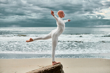 Young hairless ballerina with alopecia in white futuristic suit dancing on sea sandy beach, metaphoric surreal scene with bald pretty teenage girl exudes confidence and unique beauty