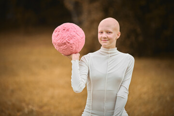 Young hairless girl with alopecia in white cloth holds pink ball in hand on fall lawn park, surreal scene with bald teenage girl gracefully interacting with symbolic pink sphere
