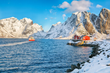 Traditional Norwegian red wooden houses on the shore of  Reinefjorden and fishing ships with  snowy mountaines in background