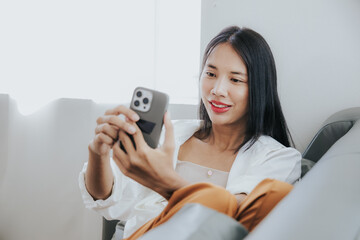 Young Asian woman sitting on a comfortable sofa at home, texting on a smartphone. Smiling young woman uses a mobile phone online message chat Buy things online from home