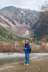Asian woman in a blue jacket standing alone, enjoying the freedom of nature. Elegant portrait of a traveler in Japan, surrounded by snow, foliage, and a serene lake.