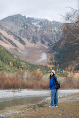 Elegant Asian woman in a blue jacket walking by a lake, surrounded by autumn foliage. A portrait of happiness and success in nature, capturing the essence of a journey.