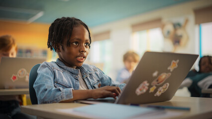 Smart Little Black Boy with Dreadlocks Sitting Behind a Desk with a Laptop Computer in Primary...