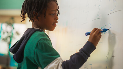 Portrait of a Handsome African Boy Finding a Solution to a Mathematical Task on a Whiteboard in Class in Elementary School. Young Pupil is Focused on Numbers, Writing Down a Correct Answer - obrazy, fototapety, plakaty