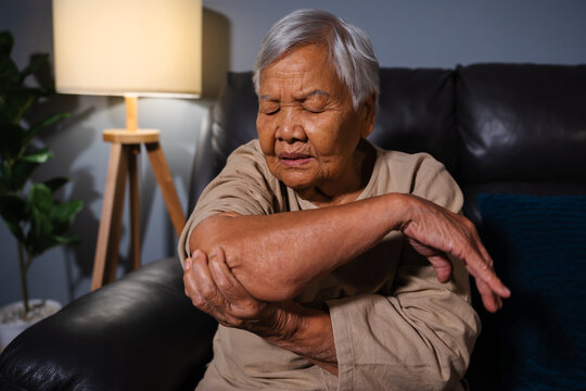 senior woman suffering from elbow pain while sitting on sofa in living room at night