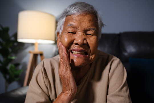 senior woman feels strong toothache while sitting on sofa in the living room at night