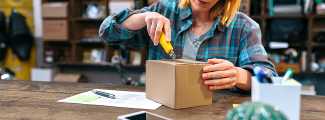 Banner of unrecognizable woman opening cardboard box with cutter in local store. Female unpacking...