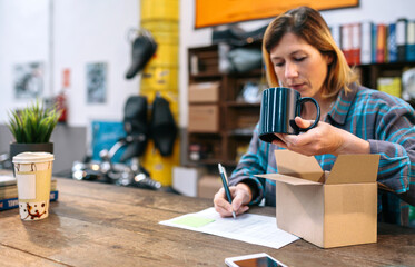 Blonde woman entrepreneur checking new merchandise received by courier to sell in her local store. Young female with freckles review mug to sent a package for client that buy in online shop.