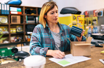 Portrait of blonde woman entrepreneur checking product received by courier to sell in her local store. Young female with freckles review blue mug to sent a package for client that buy in online shop.