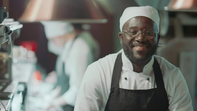 Portrait of joyous Black chef wearing apron and hat looking at camera and happily smiling at work in restaurant kitchenPortrait of joyous Black chef wearing apron and hat looking at camera and happily