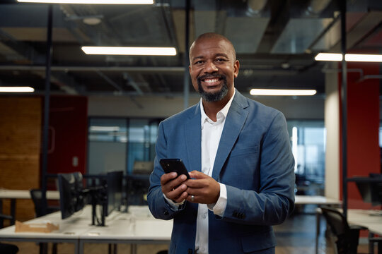 Happy mature black man in businesswear looking at camera while using mobile phone in office