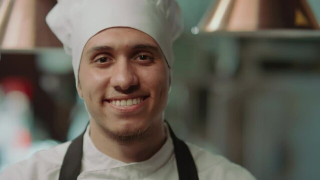 Portrait of young happy chef wearing hat and apron smiling and looking at camera while posing at work in restaurant kitchenPortrait of young happy chef wearing hat and apron smiling and looking at cam