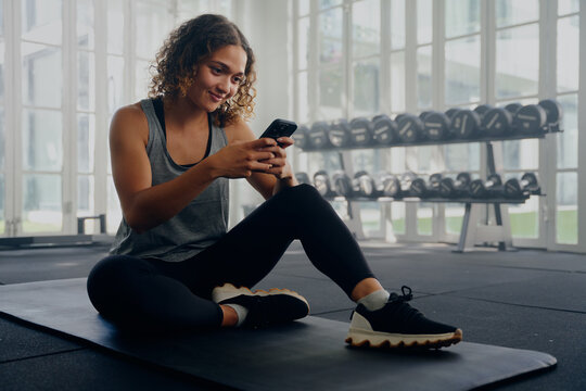 Young multiracial woman in sportswear sitting and using mobile phone while taking a break at the gym