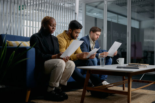 Three multiracial young adults in businesswear sitting on sofa and reading documents in office