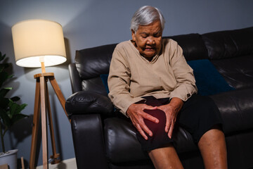 senior woman suffering from knee pain while sitting on sofa in living room at night