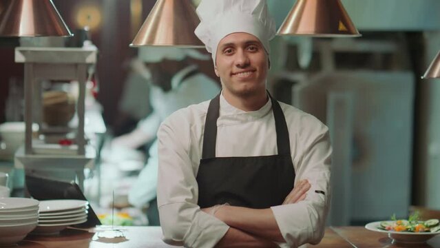 Portrait of cheerful male chef in apron and hat holding arms crossed and smiling for camera during workday in restaurant kitchenPortrait of cheerful male chef in apron and hat holding arms crossed and