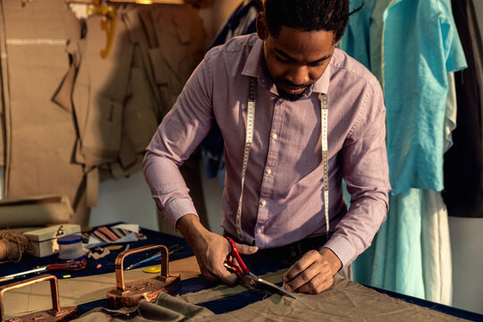 Male fashion tailor working on the suit design in his workshop cutting fabric with scissors.