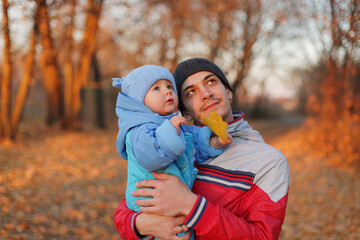 Dreamy young father embraces his baby daughter as they stroll through vibrant yellow-orange park the family reveling in outdoor play on sunny autumn day looking away