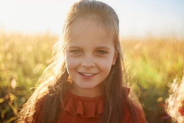 Little Caucasian joyful girl in summer field enjoying summer sunset looking smiling at camera having fun outdoor expressing positive emotions and happiness