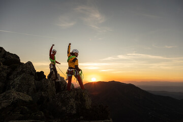 Two people are standing on a mountain, one of them is holding a camera. The sun is setting in the background, creating a beautiful and serene atmosphere